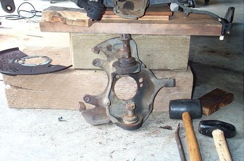 1995 Ford Bronco - A removed steering knuckle/spindle standing on a garage floor with its upper and lower ball joints still installed, propped against wood bl…