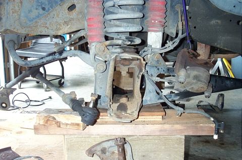 1995 Ford Bronco - Front suspension torn down with the wheel and axle shaft removed, showing the coil spring, red-booted shock, tie rod end, brake hose and st…