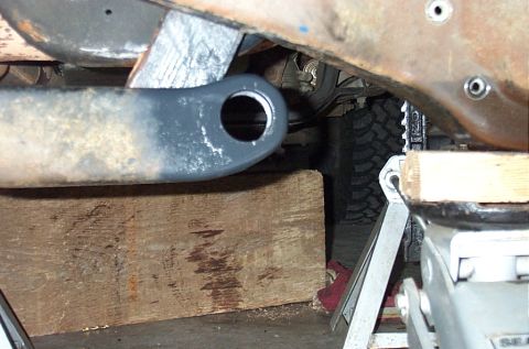 1995 Ford Bronco - Tight under-vehicle shot of a repainted gray radius arm with its bushing eye empty, resting on a wood cribbing block beneath the rusty fram…