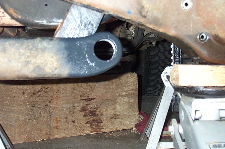Gray-painted radius arm with empty bushing eye resting on wood cribbing under a rusty truck frame