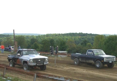 1979 Ford F-150 - Two pickup trucks compete on a dirt track, with a wooded hillside in the background and a watermarked website at the bottom.