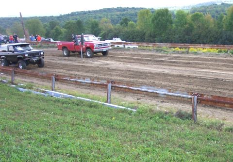 1979 Ford F-150 - Two pickup trucks compete in a race on a dirt track with spectators in the background.