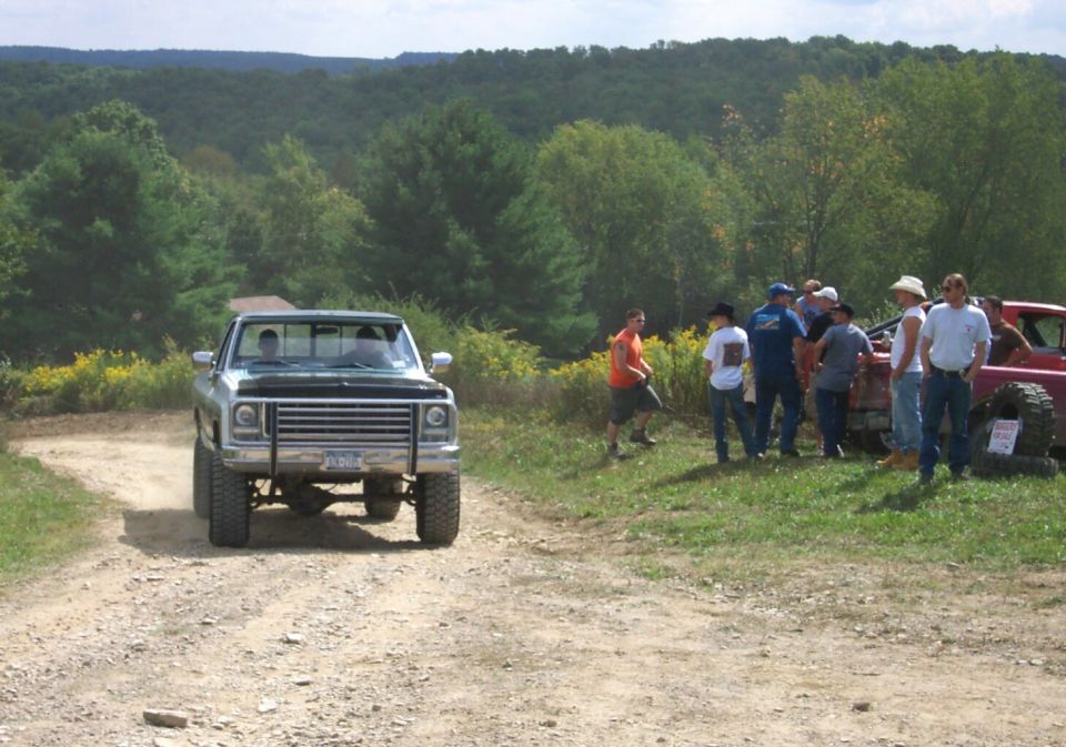 Pickup truck driving on a dirt road with a group of people standing by another vehicle in the background