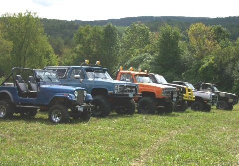 1979 Ford F-150 - Several modified off-road trucks are lined up on a grassy field with trees in the background.