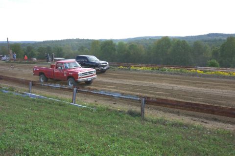 1979 Ford F-150 - Two trucks race down a dirt track, with trees and hills in the background.