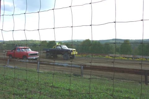 1979 Ford F-150 - Two trucks are on a dirt track with trees and hills in the background, viewed through a fence.