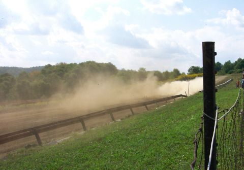 1979 Ford F-150 - A truck races along a dirt track, creating a large dust cloud. The scene is viewed from the side, with a grassy embankment and trees in the…