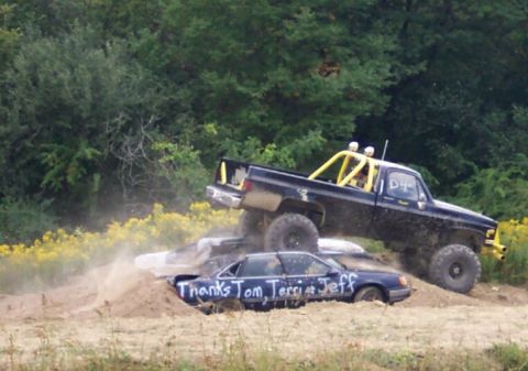 1979 Ford F-150 - A lifted pickup truck is crushing a sedan in a dirt area with trees in the background.