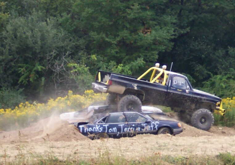 Lifted black pickup truck with yellow roll cage crushing a blue sedan. Text on sedan reads 'Thanks Tom, Terri & Jeff'
