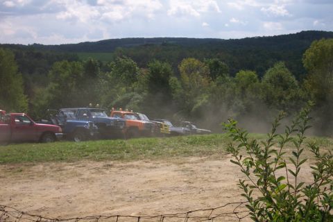 1979 Ford F-150 - A line of modified trucks and SUVs are parked on a dirt and grass embankment, with dust clouds rising around them.