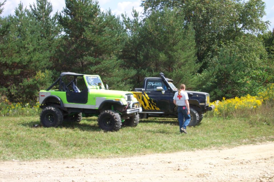 Modified green Jeep and black pickup truck parked on grassy terrain with trees in background. Text on truck reads '776-4457'