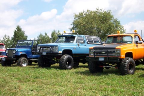 1979 Ford F-150 - Three lifted trucks, including an orange one with a black hood and a blue one, are parked on a grassy field under a partly cloudy sky.