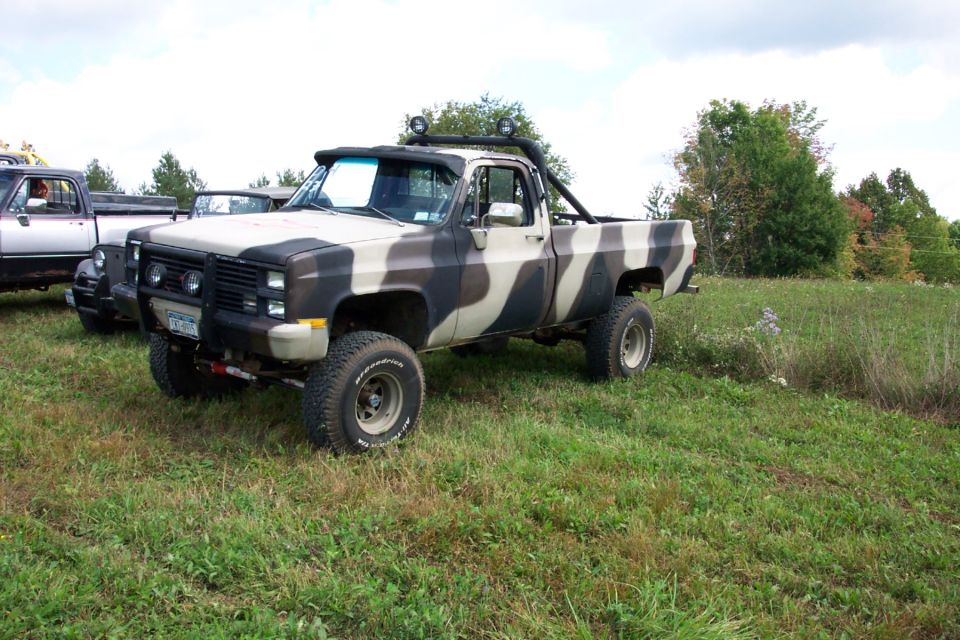 Camouflaged pickup truck with roll bar and brush guard parked in a grassy field. Another truck is visible to the left