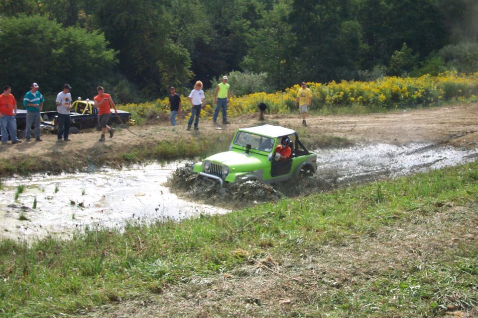 Green Jeep splashing through mud puddle, spectators watch from grassy bank