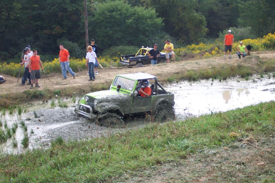 Green Jeep partially submerged in mud, with people watching and another truck in the background