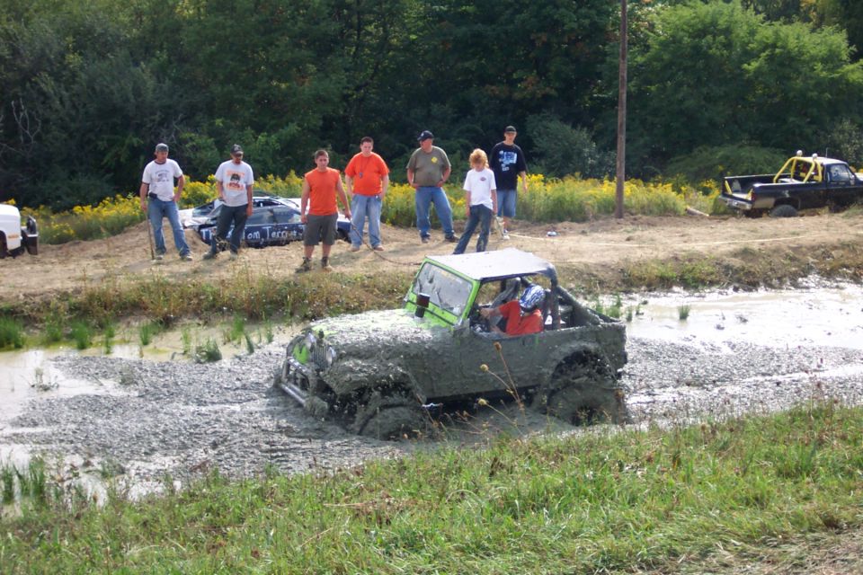 Muddy jeep driving through a water and gravel obstacle course with spectators watching