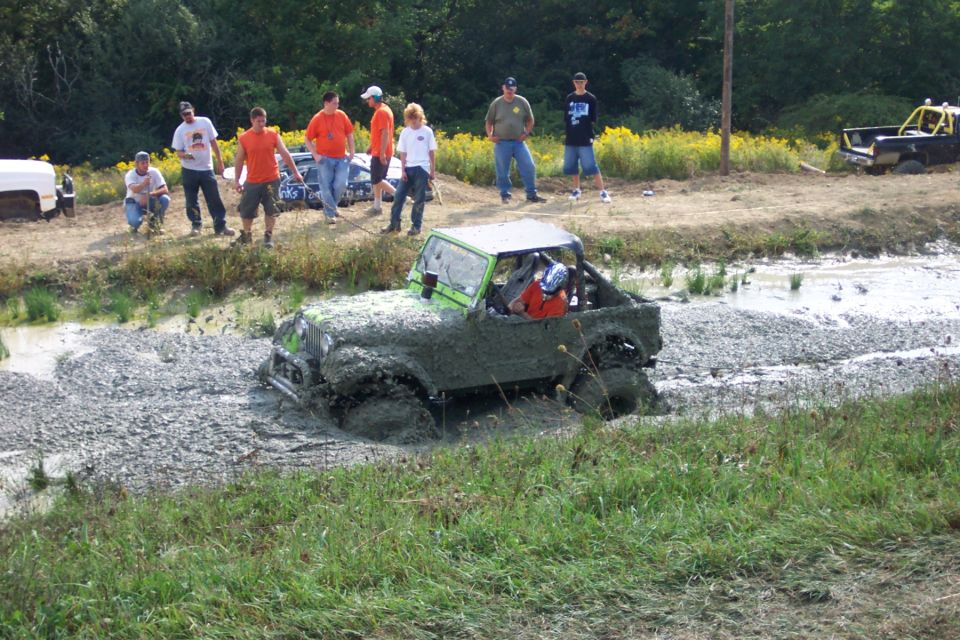 Mud-covered green off-road vehicle with a driver in an orange shirt, stuck in a muddy, rocky ditch
