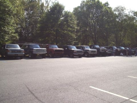 1989 Ford Bronco - A row of Ford Broncos, likely from the late 1980s, are parked in a lot during an outdoor gathering.