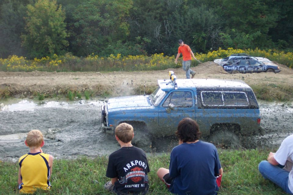 Blue SUV stuck in mud pit with people watching. Another car is in the background