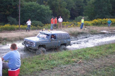 1979 Ford F-150 - A blue Chevrolet K5 Blazer navigates a muddy track, its tires deep in the mire as onlookers watch from the side.