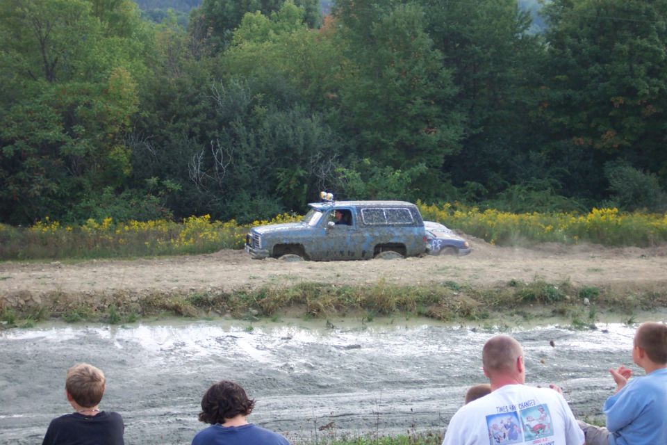 Mud bogging scene with a blue SUV and a car stuck in mud, spectators watching
