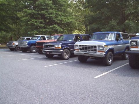 1989 Ford Bronco - Multiple generations of Ford Broncos are lined up in a parking lot on a sunny day.
