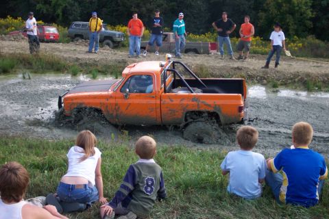 1979 Ford F-150 - Spectators watch as an orange pickup truck gets stuck in a mud pit during an off-road event.