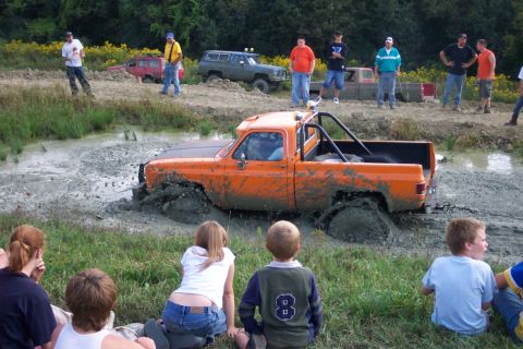 1979 Ford F-150 - An orange pickup truck is stuck deep in the mud while spectators watch.