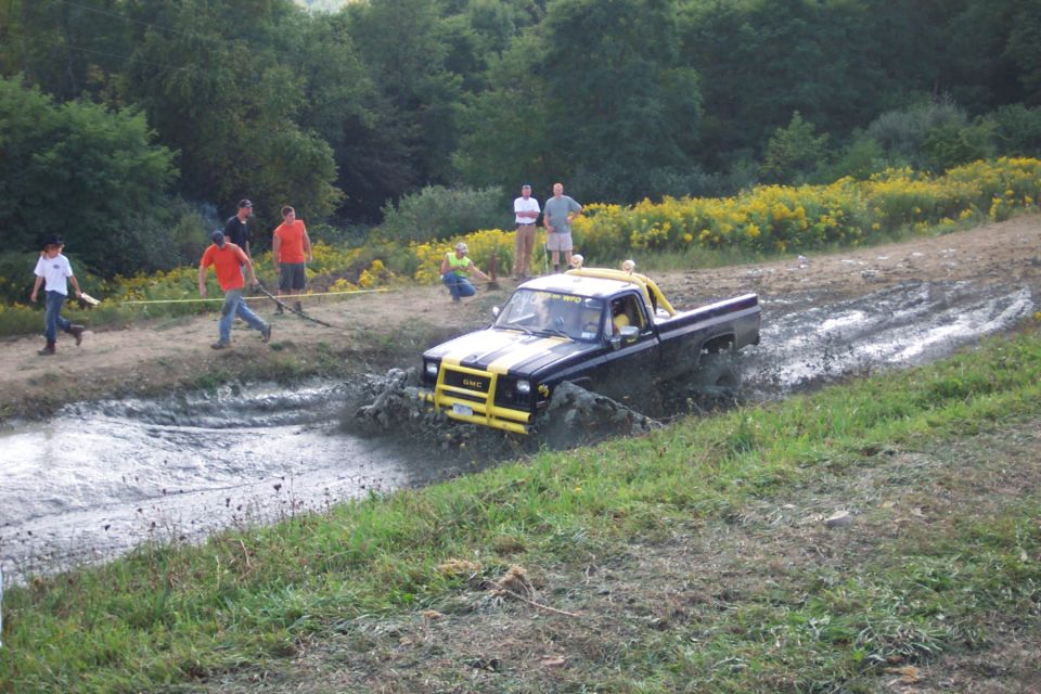 Black pickup truck with yellow stripes driving through mud, people watching on the bank