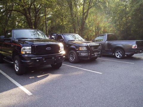 1989 Ford Bronco - Three Ford trucks are parked in a lot on a sunny day.