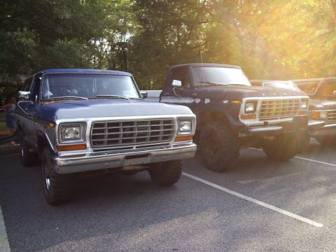 1989 Ford Bronco - Two vintage Ford trucks are parked in a lot, one blue and one dark, with trees in the background.