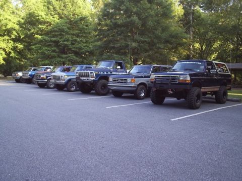 1989 Ford Bronco - Several Ford trucks and Broncos are parked in a lot, with trees in the background.