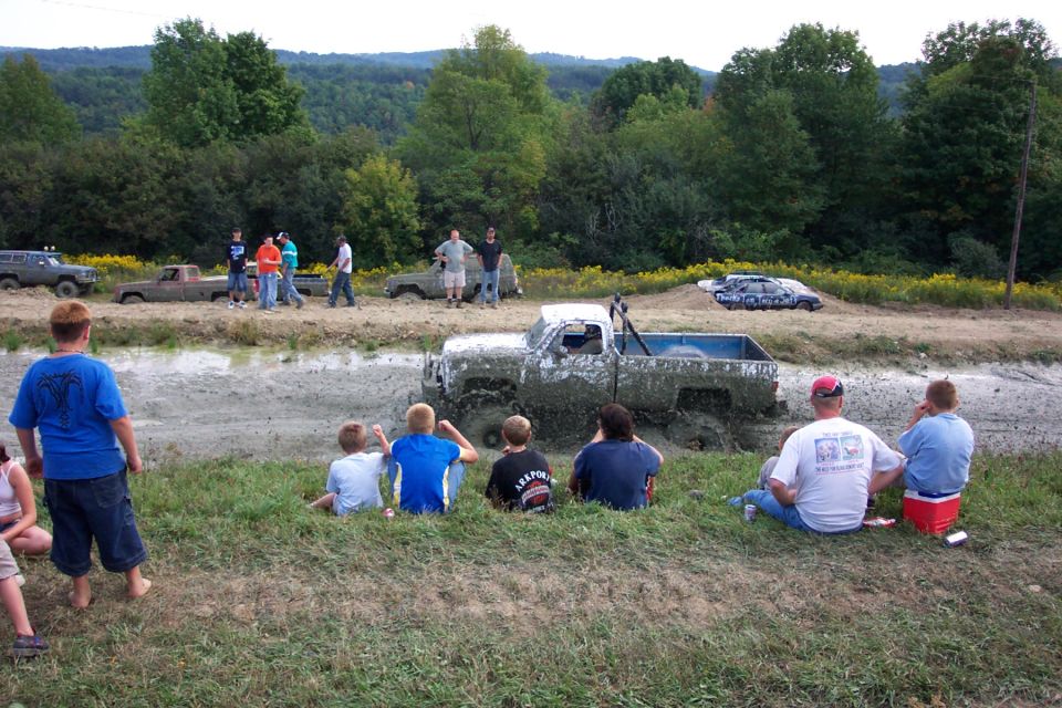Mud-covered pickup truck stuck in a mud pit, spectators watch from grassy bank. Other vehicles visible in background