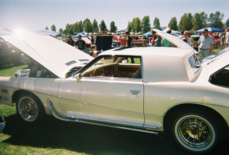 white coupe with hood open, wire wheels, and chrome trim. Other cars and people in background at an outdoor event