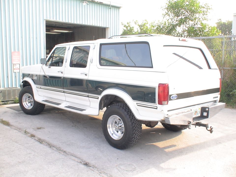 White Ford crew-cab Centurion SUV conversion with camper shell parked at a shop, rear three-quarter view