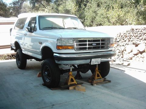 1978 Ford Bronco - A white Ford Bronco is elevated on jack stands in a driveway, showing its undercarriage.