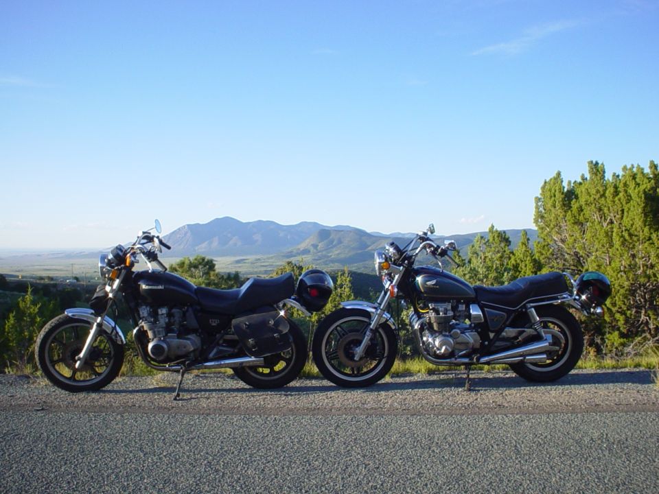 Two black motorcycles parked side-by-side on a roadside with mountains and trees in the background