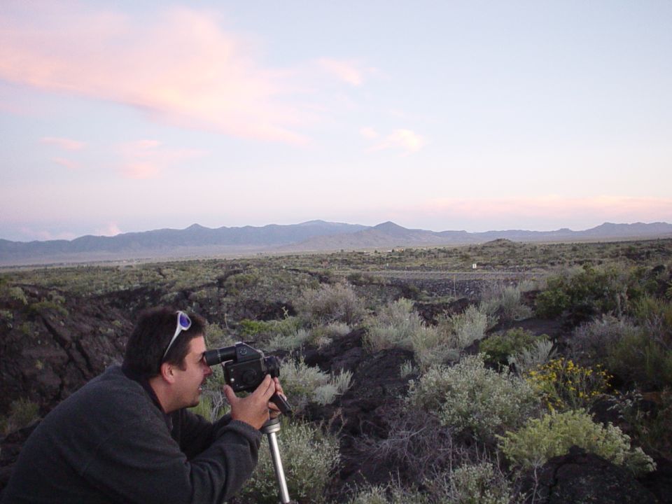 Man filming landscape with camera on tripod, mountains in background, sunset sky