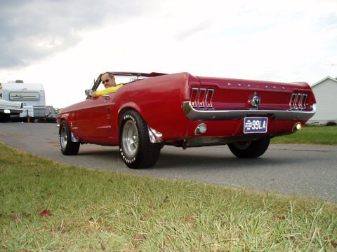 1967 Ford Mustang - A red 1967 Ford Mustang convertible is seen from the rear, driving on a road with a trailer and house nearby.