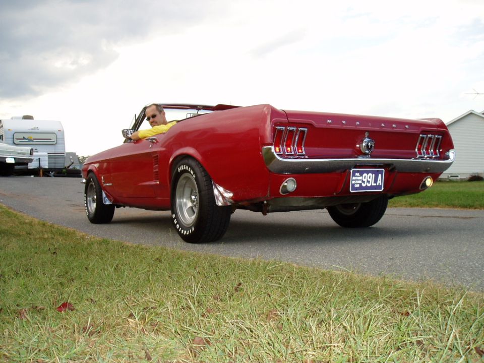 Red 1967 Ford Mustang convertible driving down a road with a trailer and house in the background