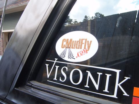 1988 Ford Bronco - A close-up view of a black SUV's rear window, two prominent stickers: 'CMudFly.com' and 'VISONIK'.