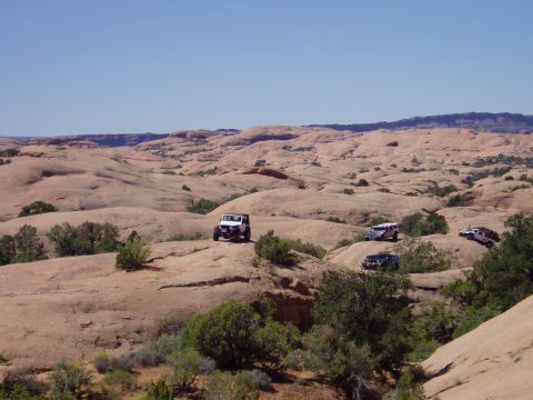 2000 Ford E350 - Wide landscape view of an off-road group spread across rolling red slickrock domes dotted with juniper. A white open-top Jeep Wrangler sits…