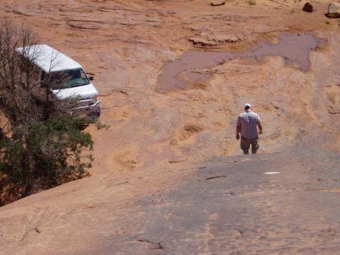2000 Ford E350 - A white full-size van sits nosed down in a red slickrock bowl at upper left, tucked beside a juniper tree with tire tracks scuffed into the…