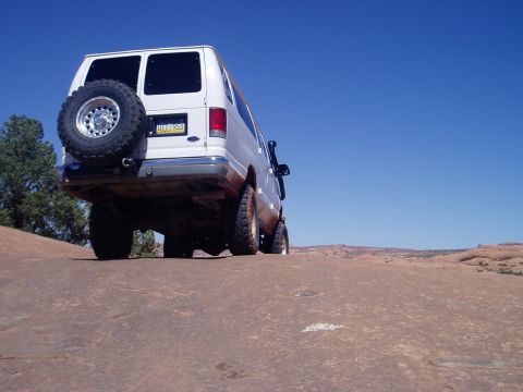 2000 Ford E350 - Low ground-level rear three-quarter shot of a lifted white Ford Econoline 4x4 van skylined on red slickrock under deep blue sky. Visible mo…