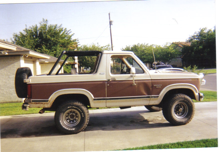 Side view of a two-tone brown and tan Ford Bronco with a spare tire on the back. It has a roll bar inside