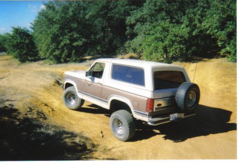 1982 Ford Bronco - This two-tone Ford Bronco is navigating a dusty, off-road trail with trees in the background.