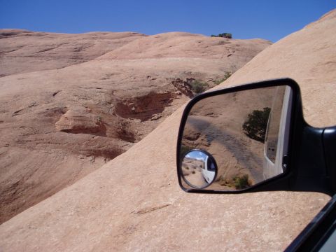 2000 Ford E350 - Shot past a black square towing-style side mirror with a stick-on convex spot mirror, looking across steeply canted red slickrock. The glas…