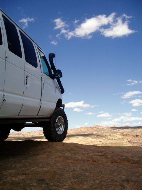 2000 Ford E350 - A white van equipped for off-roading sits on a rocky outcrop under a blue sky with scattered clouds.