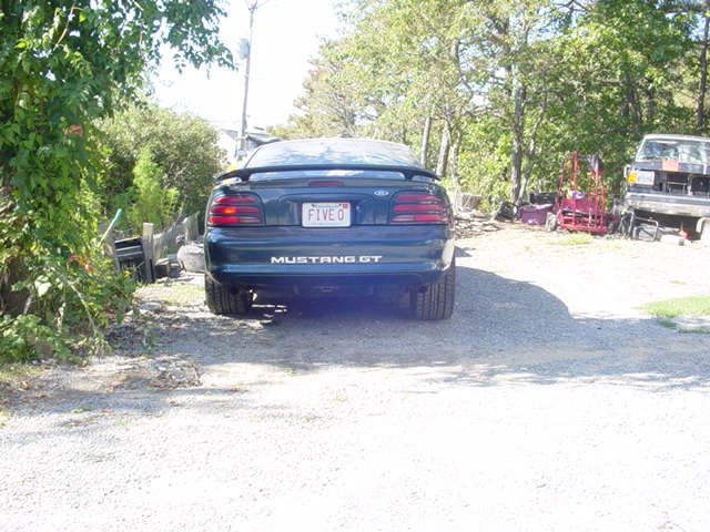 Dark green 1995 Ford Mustang GT parked outside, rear view. License plate says FIVE O. Another vehicle and some equipment are in the background