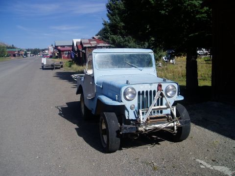 2000 Ford E350 - A light blue jeep sits parked on a gravel road in front of old buildings under a clear blue sky.